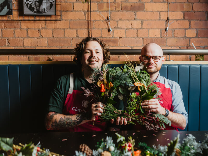 Two men proudly hold up their handmade wreath made in a Christmas craft class.